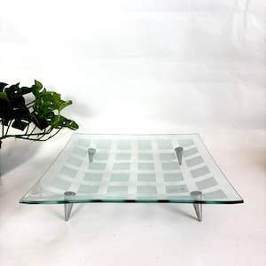 May include: Square glass serving tray with a grid pattern and four metal legs. The tray is clear with a frosted white grid design. The metal legs are silver and angled.