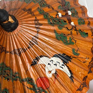 Puede incluir: Parasol de papel naranja con mango negro. El parasol presenta una grulla, un sol rojo y dise&ntilde;os de &aacute;rboles verdes y negros. Los bordes tienen un patr&oacute;n negro decorativo.