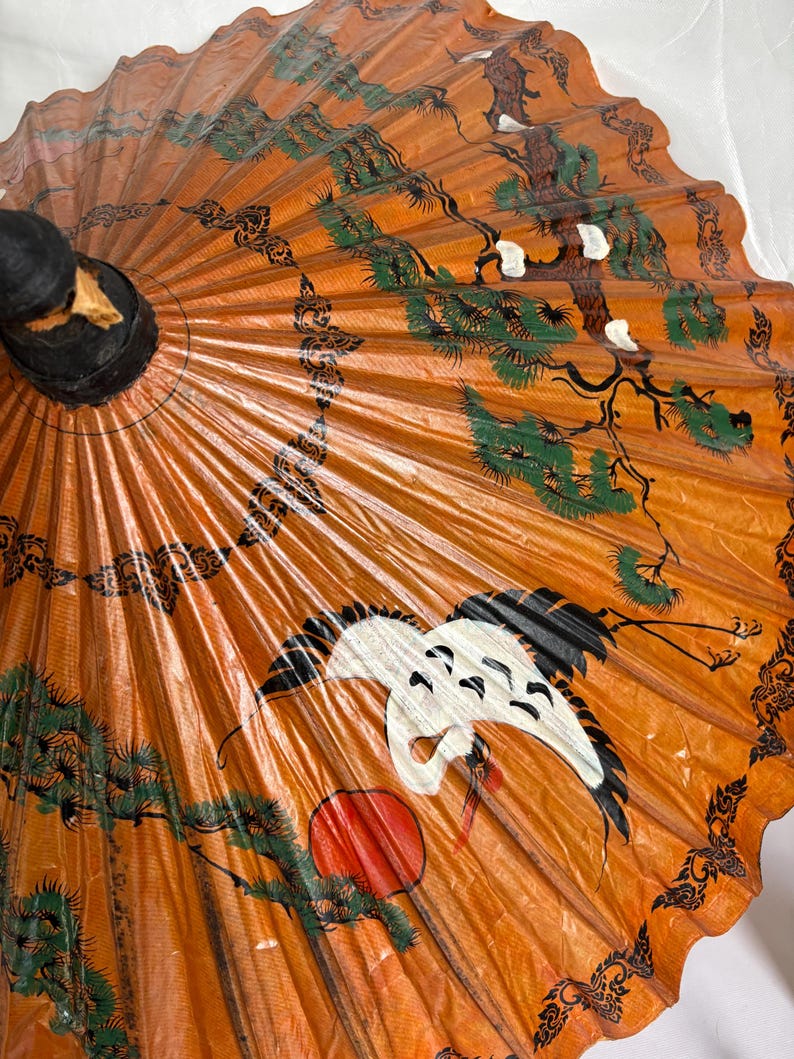 Puede incluir: Parasol de papel naranja con mango negro. El parasol presenta una grulla, un sol rojo y dise&ntilde;os de &aacute;rboles verdes y negros. Los bordes tienen un patr&oacute;n negro decorativo.