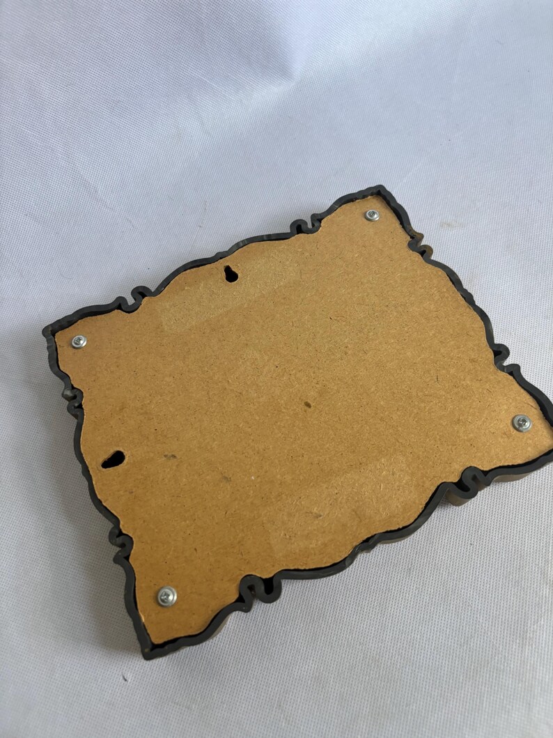 Puede incluir: Un adorno de pared decorativo con un marco ornamental marr&oacute;n oscuro. La parte posterior es de madera marr&oacute;n claro y tiene dos agujeros para colgar. Cuatro tornillos aseguran el marco al respaldo de madera.