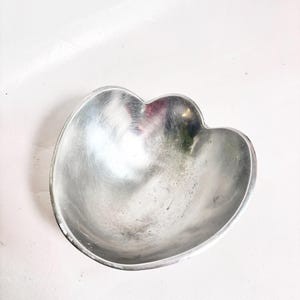 May include: A silver-coloured metal bowl with a distinct three-lobed, petal-like shape. The interior surface of the bowl features a brushed metallic finish with visible circular grain patterns and subtle variations in the reflective sheen.