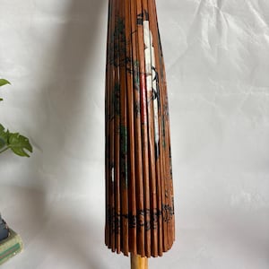 Puede incluir: Un parasol japon&eacute;s vintage cerrado. Las costillas de madera est&aacute;n cubiertas de papel marr&oacute;n con un dise&ntilde;o pintado de una grulla, &aacute;rboles y otros elementos. El mango es de madera clara.