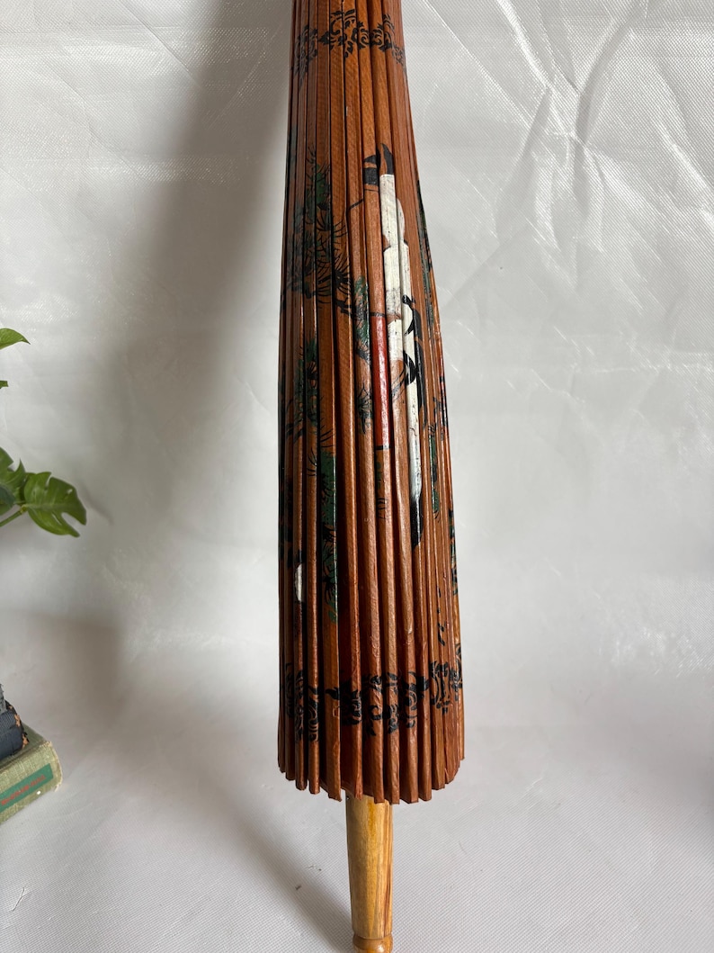 Puede incluir: Un parasol japon&eacute;s vintage cerrado. Las costillas de madera est&aacute;n cubiertas de papel marr&oacute;n con un dise&ntilde;o pintado de una grulla, &aacute;rboles y otros elementos. El mango es de madera clara.