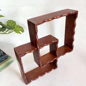 May include: A dark brown, wooden wall shelf with a unique, geometric design. The shelf features three open compartments with scalloped edges. The wood has a warm tone and visible grain, suggesting a vintage or handcrafted aesthetic.