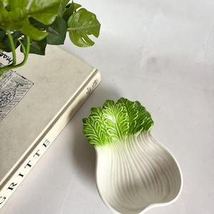 May include: A ceramic dish shaped like a stylized head of lettuce. The dish is primarily white with green detailing on the leaves. The dish is on a white surface, next to a book and a green plant.