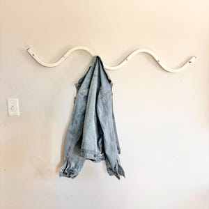 May include: A white, wavy wall-mounted coat rack with a light blue denim jacket hanging from it. The rack has a minimalist design and is mounted on a light-colored wall. The jacket is a classic style.