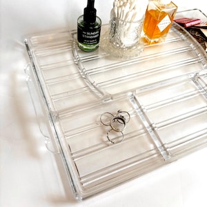 Puede incluir: Una bandeja de plástico transparente con compartimentos para organizar objetos pequeños. La bandeja tiene un borde elevado y un asa en un lado. Hay varios anillos en un compartimento, una botella de aceite esencial en otro y un frasco de vidrio con bastoncillos de algodón en un tercer compartimento.