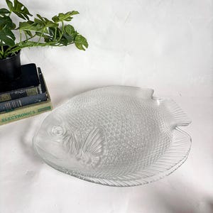 May include: A clear glass serving platter in the shape of a fish. The platter has detailed scales and fins, with a textured surface. The fish-shaped dish is a decorative piece for serving food.