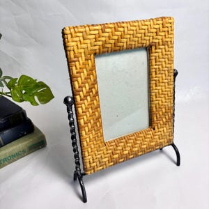 Peut inclure: Cadre photo rectangulaire avec une bordure en rotin tissé brun clair. Le cadre est soutenu par des pieds en métal noir et un support décoratif en métal torsadé. Le cadre est conçu pour tenir debout sur une surface plane.