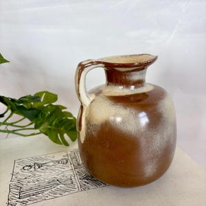 Könnte beinhalten: Ein brauner und beigefarbener Keramikkrug mit Henkel. Der Krug hat einen abgerundeten Körper und einen leicht ausgestellten Rand. Der Henkel ist gebogen und an der Seite des Kruges befestigt. Die Farben sind ein Farbverlauf, mit dunklerem Braun an der Basis und hellerem Beige oben.