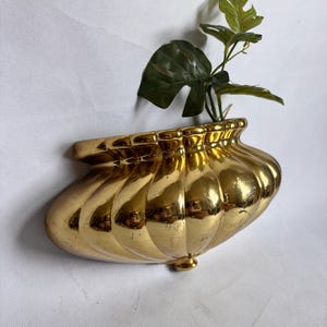 Könnte beinhalten: Ein goldfarbener Wandpflanzkübel mit gewelltem, geripptem Design und flacher Rückseite. Die metallische Oberfläche hat ein reflektierendes Finish mit sichtbaren Lichtreflexionen. Eine kleine grüne Pflanze mit mehreren Blättern steht im Gefäß.