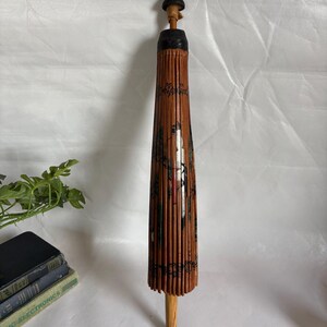 Puede incluir: Un parasol japon&eacute;s vintage cerrado, con un mango de madera y una estructura de bamb&uacute;. El parasol es principalmente marr&oacute;n con detalles pintados en negro y rojo. La parte superior es negra y el mango es marr&oacute;n claro.