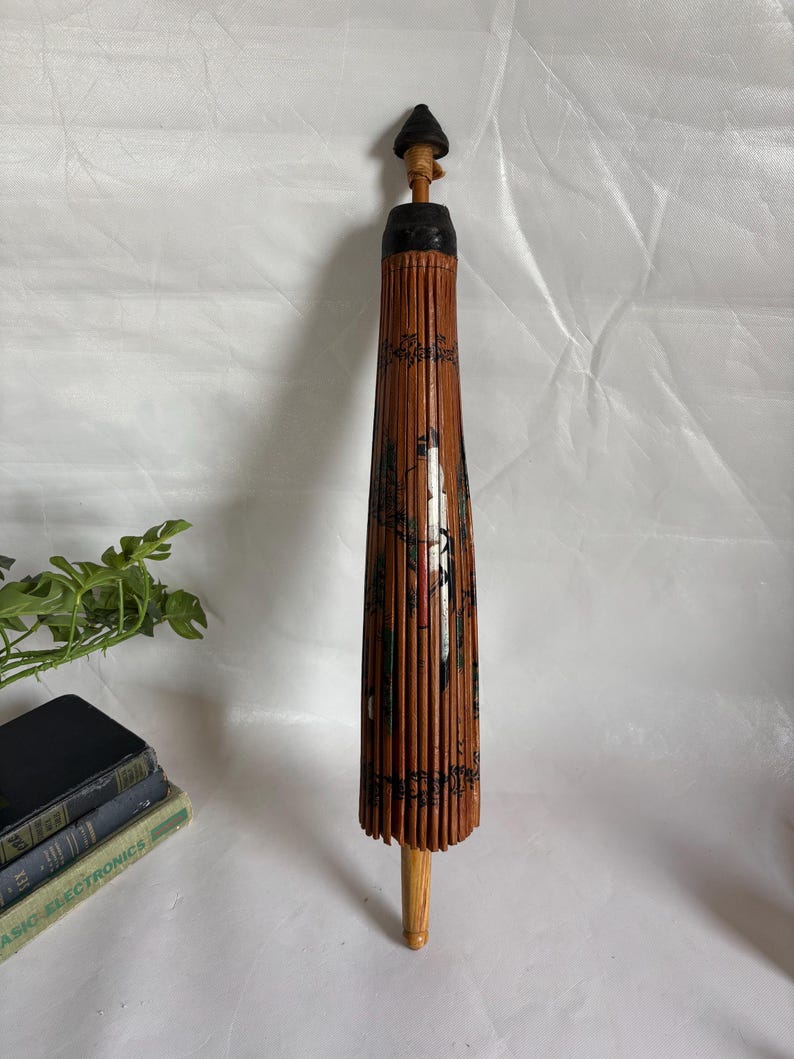 Puede incluir: Un parasol japon&eacute;s vintage cerrado, con un mango de madera y una estructura de bamb&uacute;. El parasol es principalmente marr&oacute;n con detalles pintados en negro y rojo. La parte superior es negra y el mango es marr&oacute;n claro.