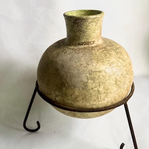 Puede incluir: Un jarrón redondo y texturizado en tonos verdes y beige, colocado en un soporte de metal oscuro. El jarrón tiene un cuello estrecho y un cuerpo bulboso. El soporte de metal tiene un anillo circular para sujetar el jarrón y tres patas curvas.