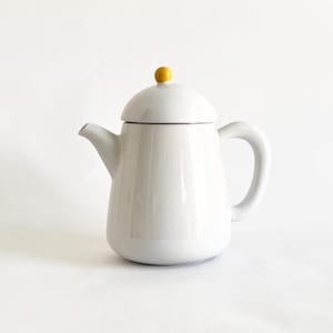 May include: A white ceramic teapot with a rounded body, a curved handle, and a spout. The lid is topped with a small, yellow knob. The teapot is designed for brewing and serving tea.