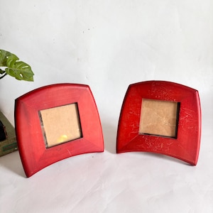 Peut inclure: Deux cadres photo rouges assortis avec un bord supérieur et inférieur incurvé. Chaque cadre présente une ouverture centrale carrée avec un fond beige. Les cadres ont une finition mate lisse et une silhouette arquée distincte, tenant debout.