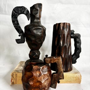 May include: Four hand-carved wooden vessels with dark brown finishes. The collection includes a tall vase, a mug, and a small cup, all with textured surfaces and unique handle designs. The items are displayed on top of a vintage book.
