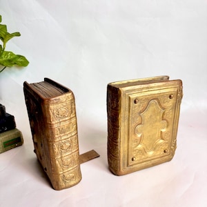 May include: Two gold-toned metal book-shaped objects with ornate embossed detailing on the covers and spines. One object is positioned upright showing the spine with horizontal ridges, while the other is lying flat displaying the front cover.