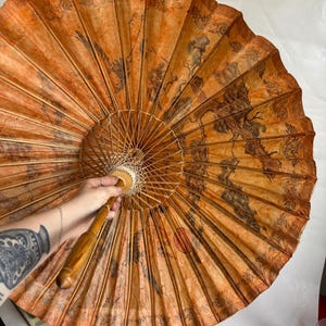Puede incluir: Un parasol redondo y abierto, de color naranja y marr&oacute;n, con un mango de madera. El parasol presenta una detallada ilustraci&oacute;n negra de un paisaje con &aacute;rboles y p&aacute;jaros. El parasol est&aacute; hecho de papel y bamb&uacute;.