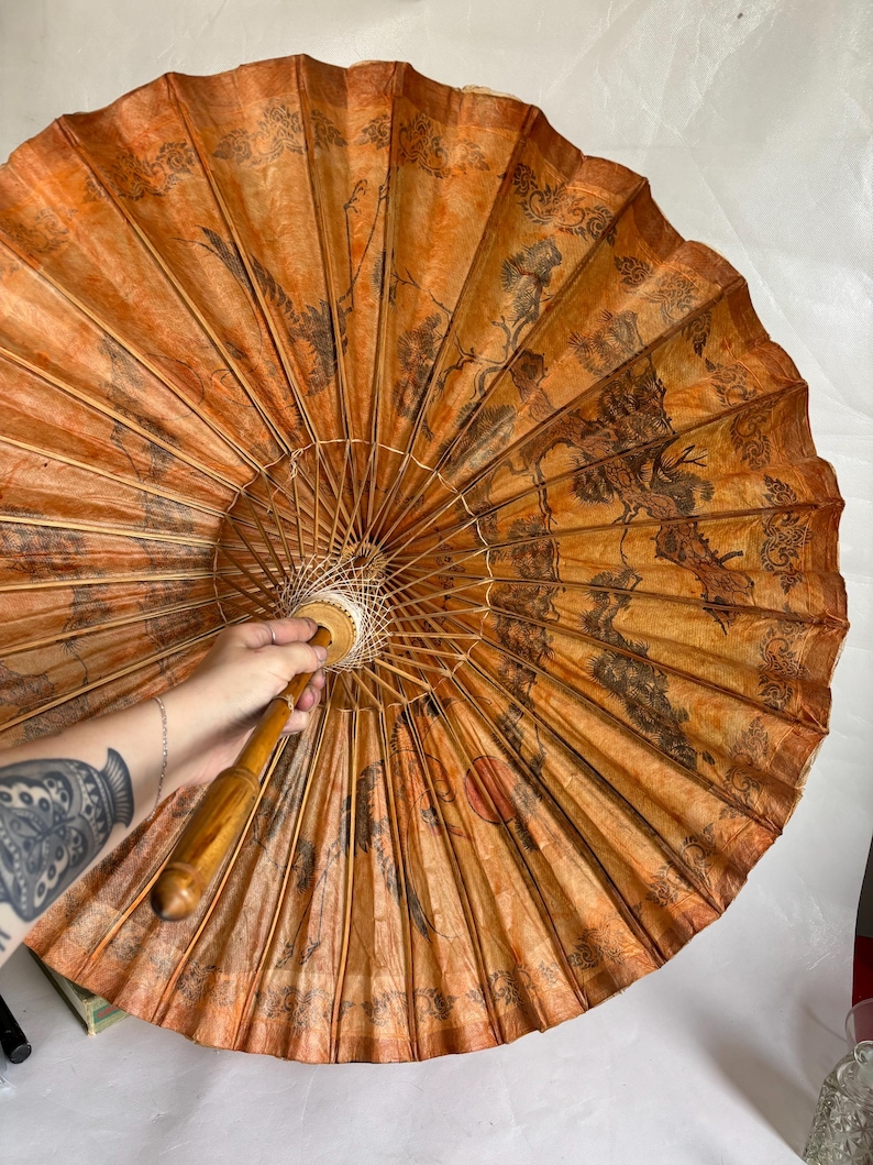 Puede incluir: Un parasol redondo y abierto, de color naranja y marr&oacute;n, con un mango de madera. El parasol presenta una detallada ilustraci&oacute;n negra de un paisaje con &aacute;rboles y p&aacute;jaros. El parasol est&aacute; hecho de papel y bamb&uacute;.