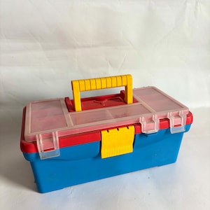 May include: A colorful plastic toolbox with a blue base, red trim, and a clear lid. The toolbox has a yellow handle and a yellow latch. The lid has several small compartments. The toolbox is designed for storing tools and small items.