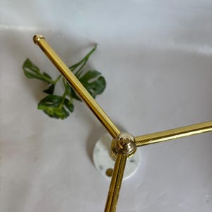 May include: A gold-colored metal decorative stand with a circular marble base and three arms extending outwards. The stand has a modern design and is suitable for displaying small items.
