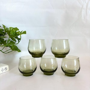 May include: A set of six olive-green stemless drinking glasses. The glasses have a rounded shape with a slightly darker base. They are arranged in a group on a white surface, suggesting a vintage or retro aesthetic.