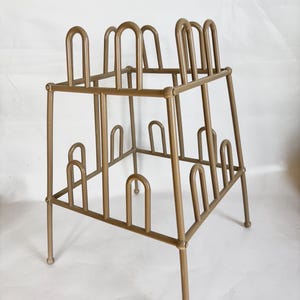May include: A vintage metal boot rack in a warm, neutral tone. The rack has a square frame with arched supports designed to hold multiple pairs of boots. The legs are slightly angled, and the overall design is simple and functional.