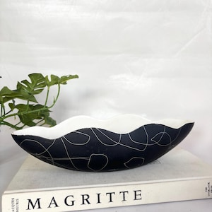 Op de afbeelding: Een zwart-witte keramische kom met een golvende rand. De kom heeft een zwarte buitenkant met witte abstracte lijn- en stipontwerpen. De binnenkant is wit. De kom ligt op een boek met de titel "MAGRITTE".