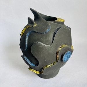 May include: A dark gray ceramic vase with an abstract design. The vase has a textured surface with yellow and blue accents. The vase has a unique shape with two pointed protrusions at the top. The vase is a decorative item.