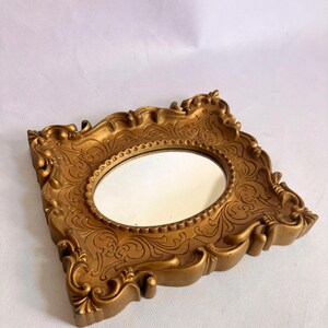 Puede incluir: Espejo dorado ornamentado con una superficie de espejo ovalada. El marco presenta detalles de volutas y adornos decorativos. El espejo es un art&iacute;culo decorativo de estilo vintage.