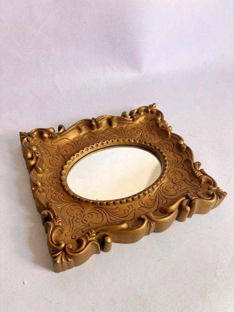 Puede incluir: Espejo dorado ornamentado con una superficie de espejo ovalada. El marco presenta detalles de volutas y adornos decorativos. El espejo es un art&iacute;culo decorativo de estilo vintage.