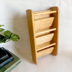 May include: A light-colored wooden magazine rack with three horizontal shelves. The rack has a rectangular shape with rounded edges and is designed to hold magazines or books. The wood grain is visible.