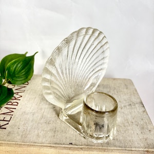 Puede incluir: Un plato de vidrio transparente en forma de concha con una pequeña taza cilíndrica de vidrio adjunta. La concha tiene una textura acanalada detallada. Los objetos están sobre un libro con la palabra "REMBRANDT" visible. Decorativo y vintage.