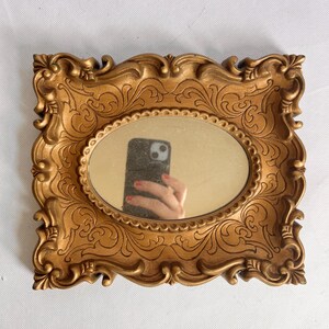 Puede incluir: Un espejo rectangular dorado con un espejo ovalado en el centro. El marco tiene un dise&ntilde;o ornamental y enrollado. El espejo refleja una mano que sostiene un tel&eacute;fono.