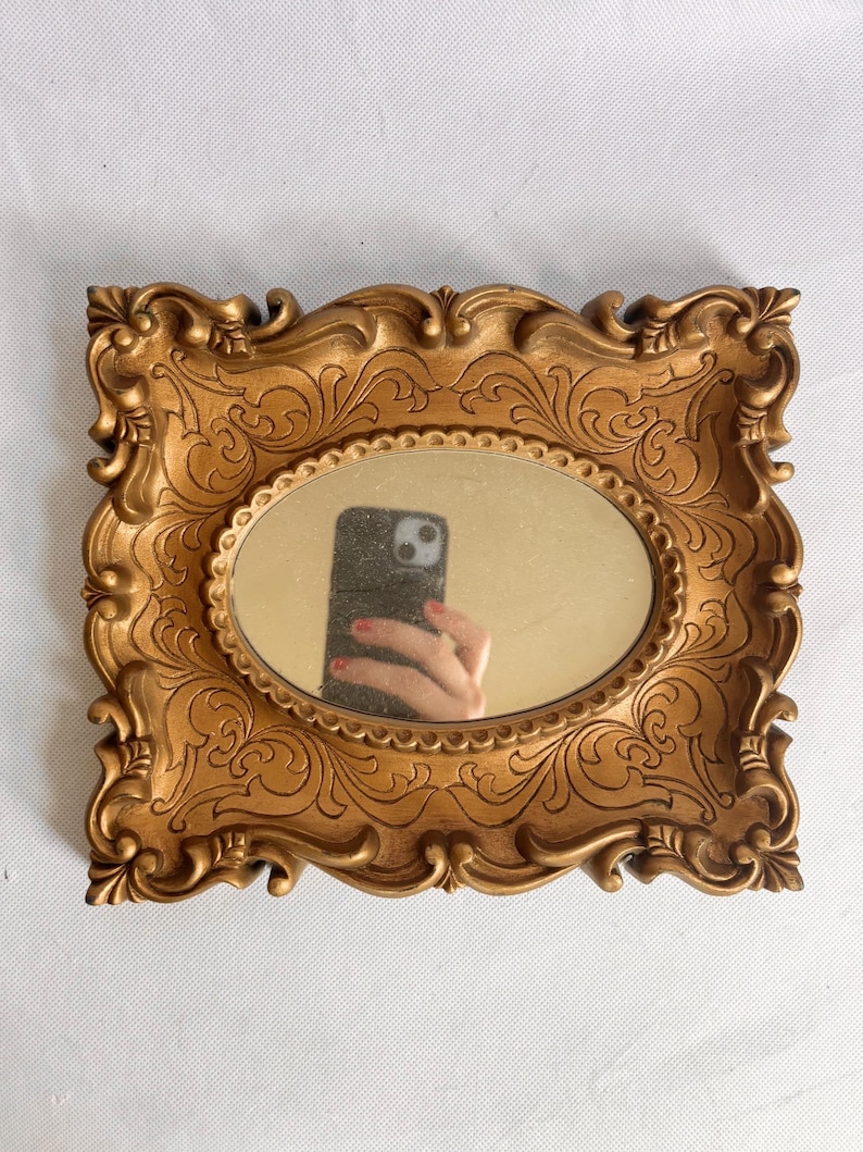 Puede incluir: Un espejo rectangular dorado con un espejo ovalado en el centro. El marco tiene un dise&ntilde;o ornamental y enrollado. El espejo refleja una mano que sostiene un tel&eacute;fono.