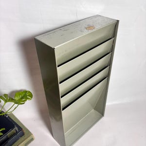 May include: A light olive green metal wall-mounted document organizer featuring five tiered slots for storage. The rectangular unit has a smooth finish with some visible wear on the top edge and a small circular label affixed to the top surface.