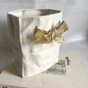 May include: A white ceramic vase with a crumpled paper bag design, adorned with a gold ribbon bow. The vase is placed on a book titled "MAGRITTE". The overall aesthetic is minimalist and elegant.