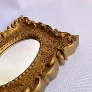 Puede incluir: Un espejo ovalado con un marco dorado y ornamentado. El marco presenta detalles de volutas y una superficie texturizada. El espejo est&aacute; en &aacute;ngulo, mostrando un reflejo. El dise&ntilde;o es vintage y decorativo.
