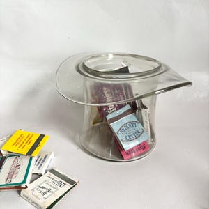 Puede incluir: Un recipiente transparente de vidrio en forma de sombrero lleno de cajas de cerillas. Varias cajas de cerillas están esparcidas alrededor del recipiente. Las cajas de cerillas tienen varios diseños y texto, incluyendo "HOLLY'S LANDING".