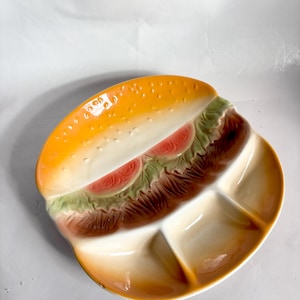 Puede incluir: Un plato de cerámica con forma de hamburguesa. Presenta un pan amarillo-naranja, lechuga verde, rodajas de tomate rojas y una hamburguesa marrón. El plato está dividido en secciones. Un plato para servir divertido y original.