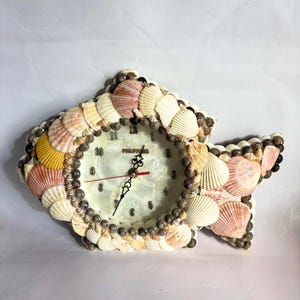 Puede incluir: Un reloj de pared con forma de pez decorado con varias conchas marinas. La esfera del reloj es blanca con números y manecillas negras, y un segundero rojo. El reloj está hecho de conchas en tonos blancos, rosas y marrones.