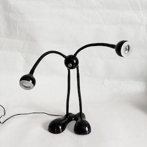 May include: A black desk lamp with three adjustable heads. The lamp has a rounded base and flexible arms, allowing for directional lighting. The lamp's design is modern and minimalist, suitable for a workspace or bedside table.