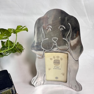 Peut inclure: Un cadre photo en métal argenté en forme de chien aux longues oreilles. Le centre présente une ouverture rectangulaire pour une photo de 10 cm x 15 cm. Le papier inséré affiche le texte "PHOTO FRAME 4"X6" 10CMX15CM" et des empreintes de pattes.