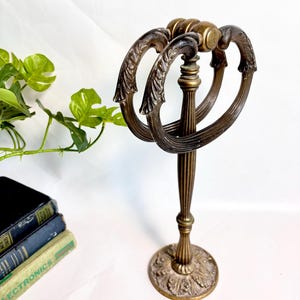 May include: An antique brass towel rack with a decorative floral base and three oval rings. The rack has a central column with vertical grooves and a decorative top. The overall height is approximately 12 inches.