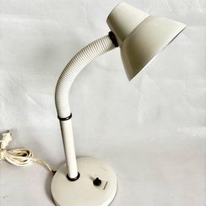 May include: A vintage off-white desk lamp with a flexible, ribbed neck and a round base. The lamp features a conical shade and a power switch on the base. The cord and plug are also off-white.