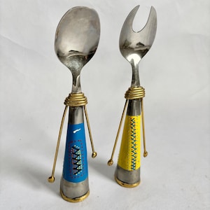 May include: A vintage serving spoon and fork set. The stainless steel utensils have decorative handles with a blue and yellow painted design. The handles are supported by brass-colored wire legs and a gold-colored band.