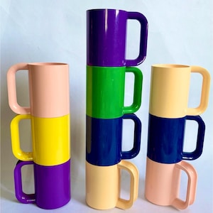 May include: Stacked colorful mugs in shades of purple, green, blue, yellow, and peach. Each mug has a large, curved handle. The mugs are arranged in three stacks, each with a different color combination.