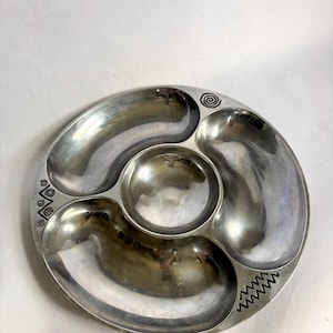 Puede incluir: Una fuente plateada para servir con cuatro compartimentos. Tiene una zona central circular rodeada de tres secciones en forma de riñón. El borde presenta patrones de espirales y zigzags grabados. Adecuada para servir aperitivos o snacks.