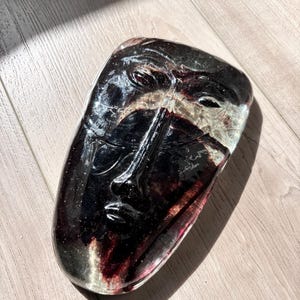 May include: A smooth, oval-shaped glass sculpture featuring a stylized human face molded in relief. The piece is crafted from deep, translucent dark red and black glass, creating a layered effect that highlights the facial features.
